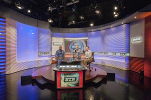BTN Broadcast Studio with illuminated blue wall backdrop, with three men sitting around an anchor desk with BTN logo