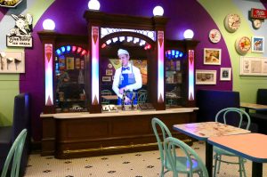 Old fashiones styled ice cream dispensary made of brass and wood with marble top fitted with a screen that plays footage of a server dressed in blue bunny apron and hat.